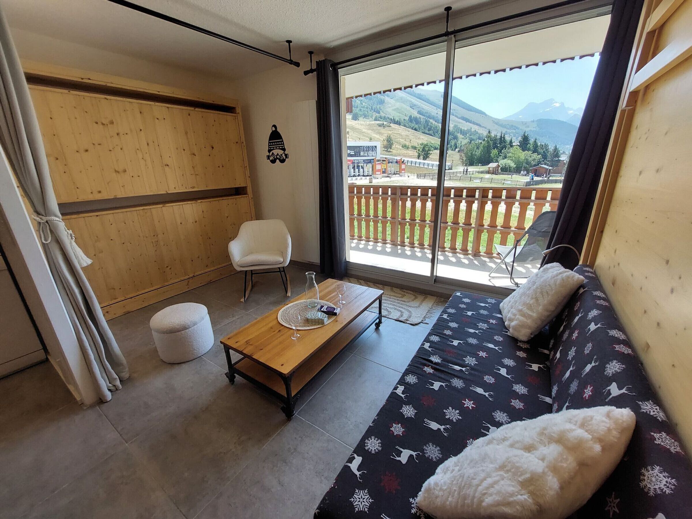 A cozy room with a wooden floor, a sofa with patterned cushions, a small table with a glass, and a chair. A large window offers a view of a balcony and mountains.