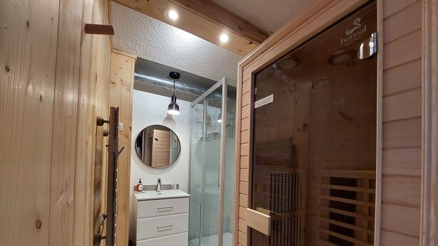 Un baño moderno con tocador de madera, un espejo redondo, cabina de ducha de vidrio y puerta a la sauna.