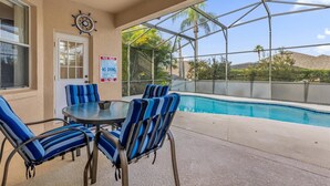 Un espace piscine avec des chaises à rayures bleues et une table, un panneau indiquant les règles à respecter au bord de la piscine, et une vue sur des palmiers et des montagnes.