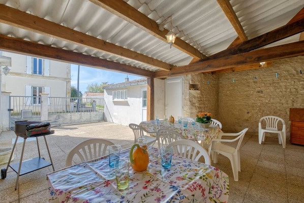 An outdoor patio with a table set for dining, surrounded by white chairs, under a corrugated metal roof.