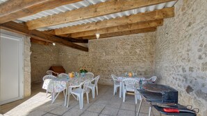 Un patio exterior con muros de piedra, vigas de madera, sillas de plástico blanco, una mesa con una parrilla y una puerta.