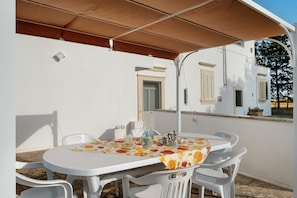 Un patio al aire libre con una mesa redonda, sillas de plástico blancas y un mantel con estampado floral.