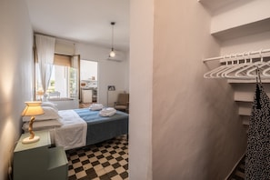 A hotel room with a bed, a lamp, a checkered floor, a window with curtains, and a hanging clothes rack.
