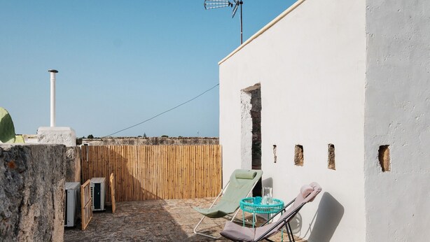 Eine Dachterrasse mit weißer Wand, einem Bambuszaun, einem grünen Lounge-Sessel und einem kleinen Tisch mit einem Kännchen.
