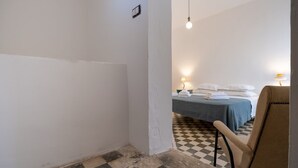 Ein Hotelzimmer mit Schachbrettmuster-Boden, einem Bett mit weißen Laken, einem Sessel und einer Wandsteckdose.