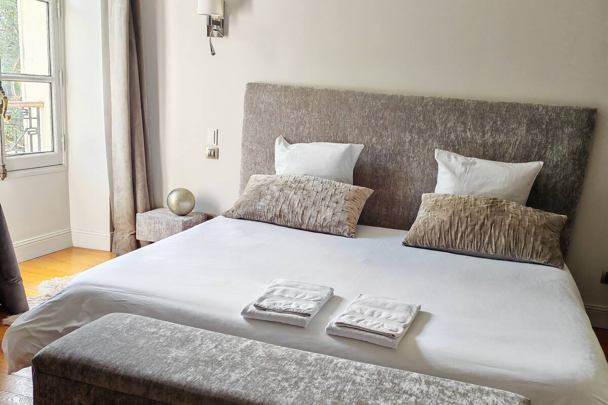 A neatly made bed with white linens, a grey upholstered bench, and a bedside table with a lamp.
