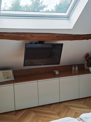 A room with a sloped ceiling, a flat-screen TV mounted on a wooden shelf, a framed certificate on the left, and a small table with two bottles and a candle. - villa raspail (Ivry-sur-Seine)