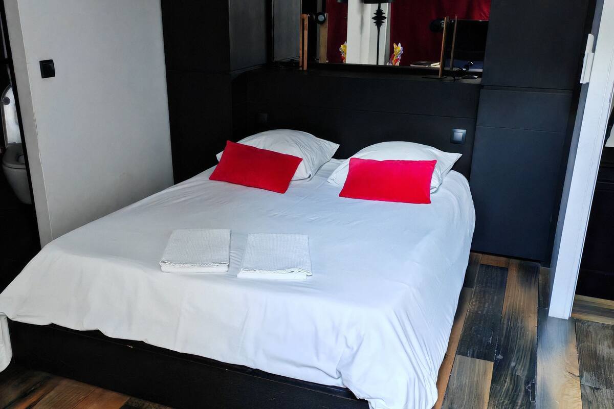 A modern bedroom with a large bed, dark wooden flooring, and a red accent pillow.