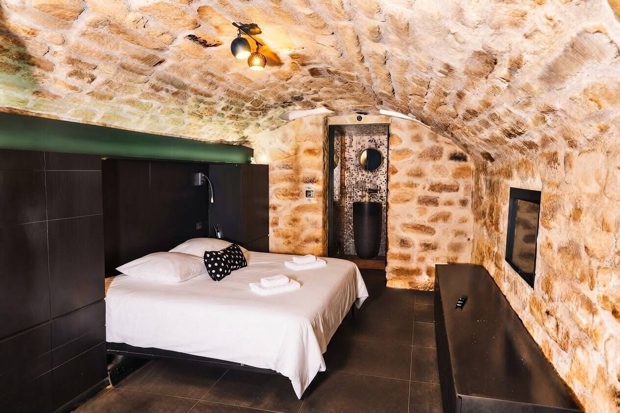 A room with a stone ceiling, a bed with white linens, a bench, and a wall-mounted TV.