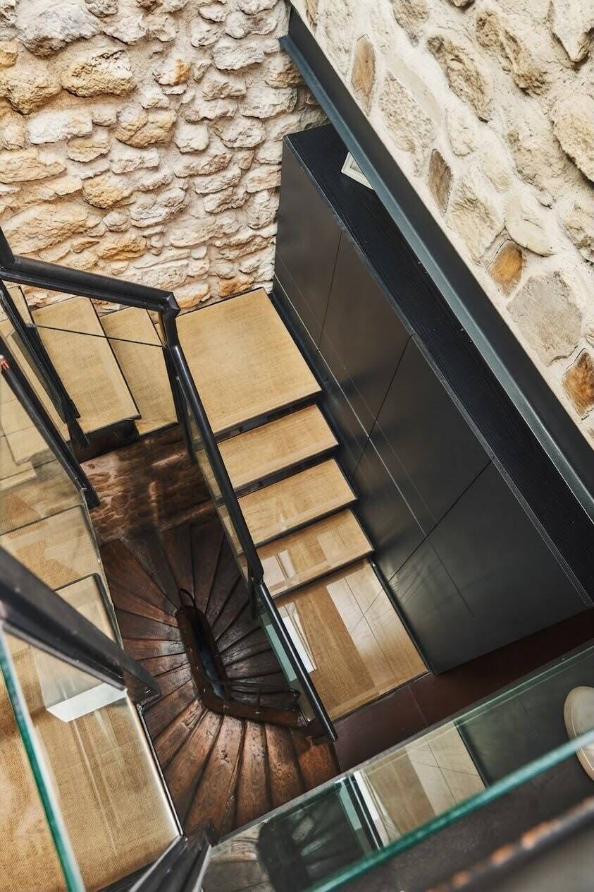 Un escalier en colimaçon avec des marches en bois et une rampe en verre, menant à une pièce dotée d’un mur en pierre et d’un meuble noir au design moderne.