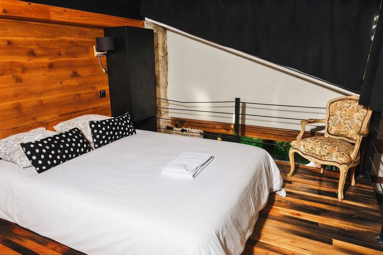 Un dormitorio con cabecera de madera, cama con ropa de cama blanca y almohadas con lunares negros, una silla con cojín estampado y una cortina negra.