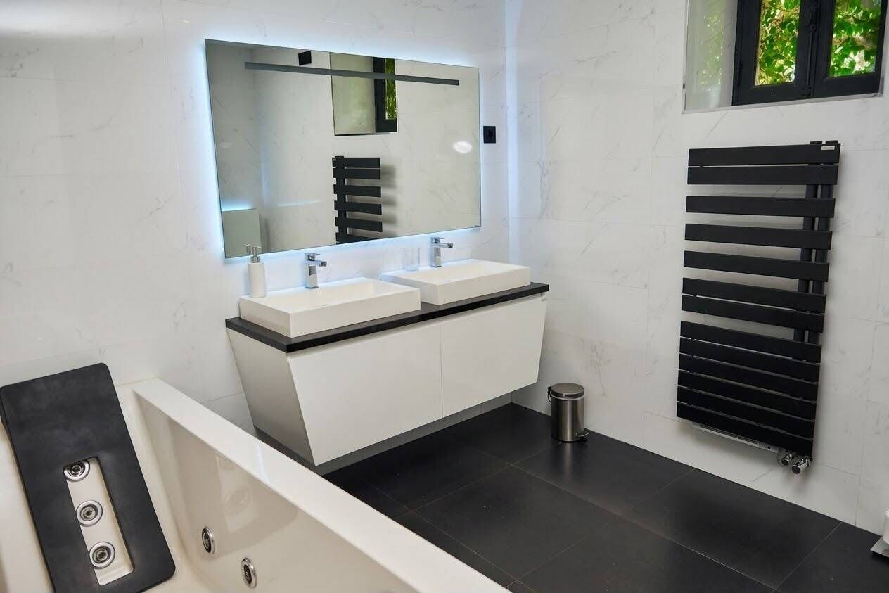 A modern bathroom with a large mirror, two sinks, a bathtub, and a black towel rack.