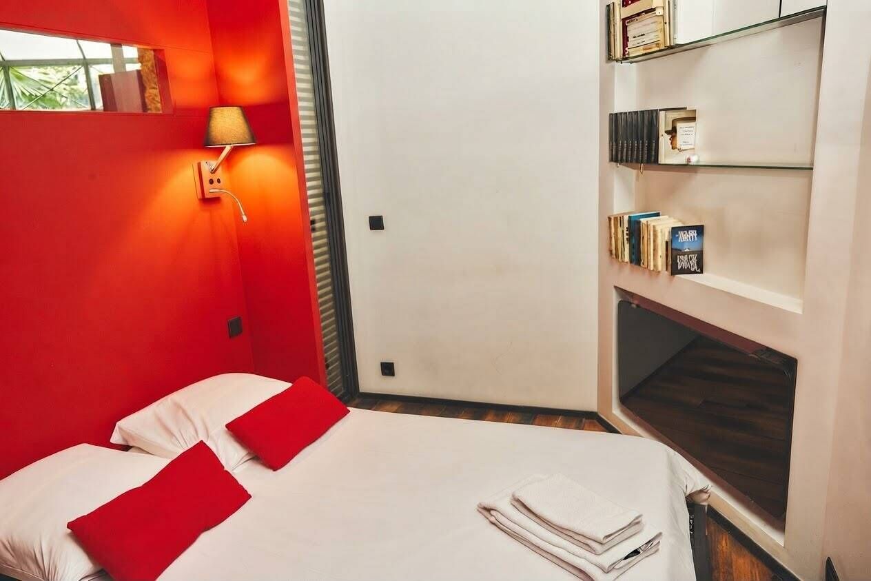 Una habitación de hotel con cama, almohadas rojas, lámpara de pared, estantería y ventana.
