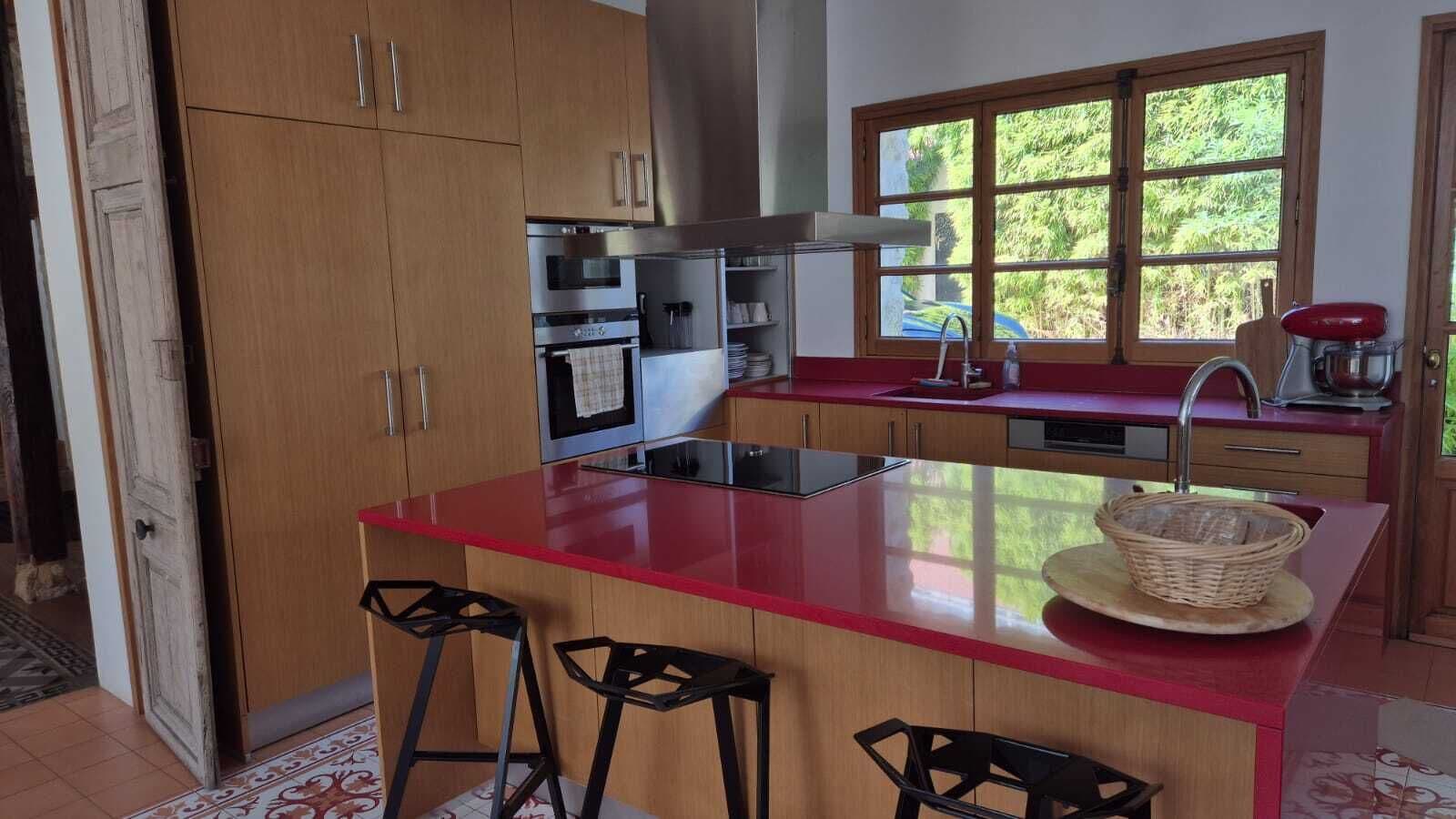 Uma cozinha com armários de madeira, bancada vermelha, forno embutido, pia e janela com vista para área verde.