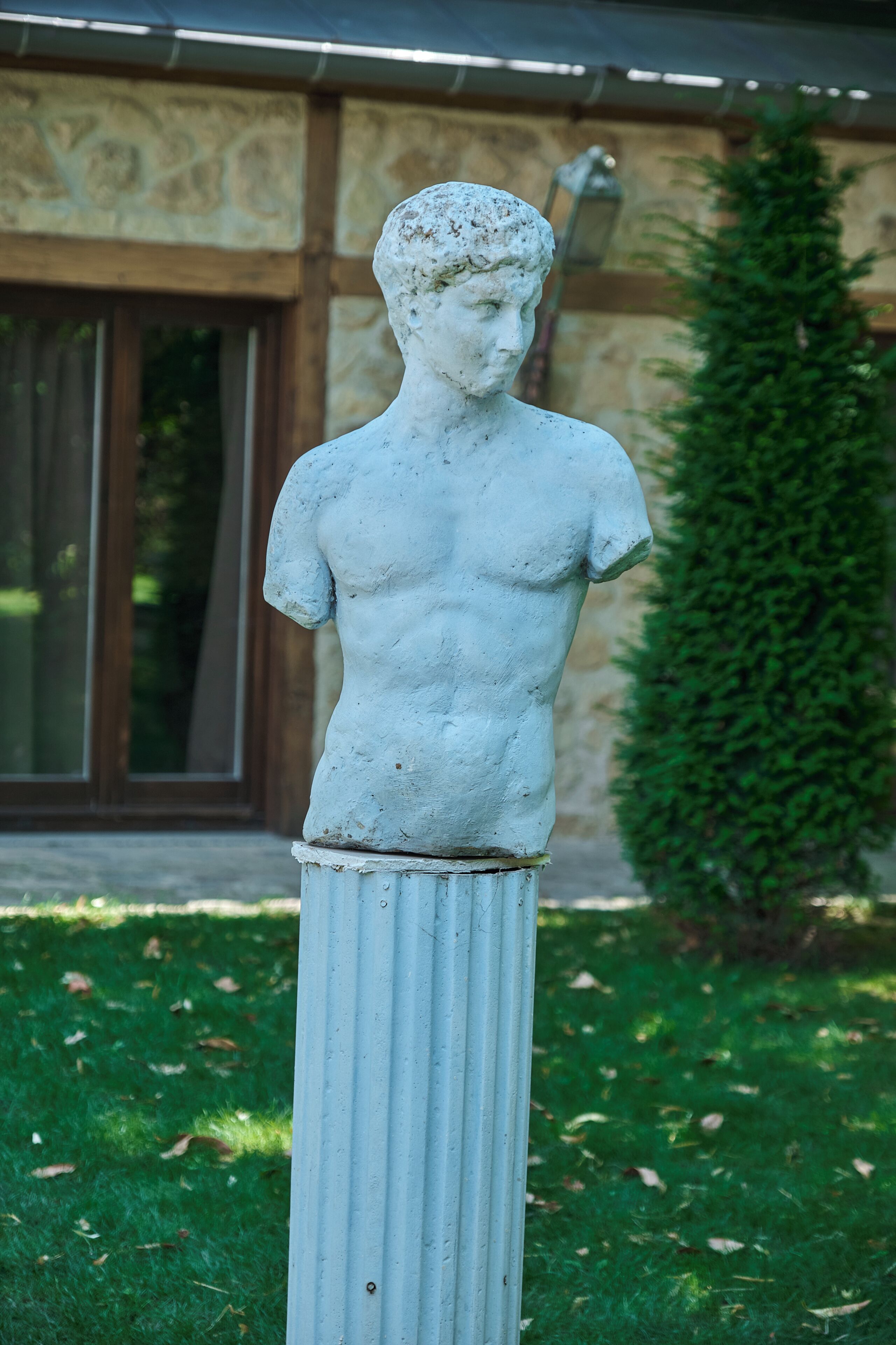 A stone statue of a human figure with a pedestal, set against a building facade and a green hedge.