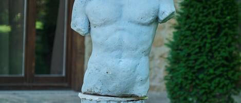 A stone statue of a human figure with a pedestal, set against a building facade and a green hedge.