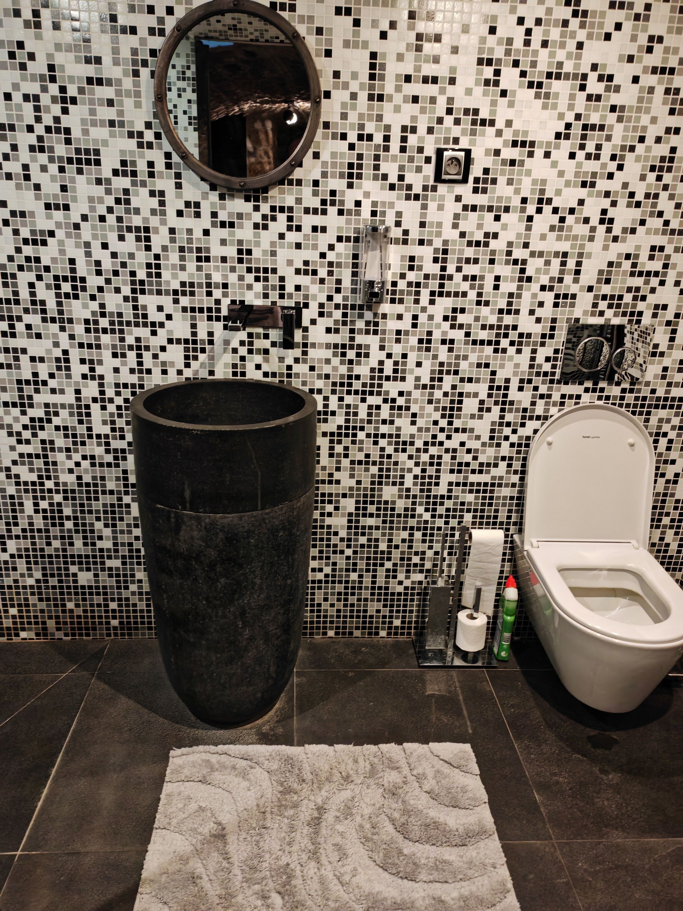 Ein Badezimmer mit Wand aus Mosaikfliesen, einem runden Spiegel, einer Toilette und einer großen schwarzen Mülltonne.
