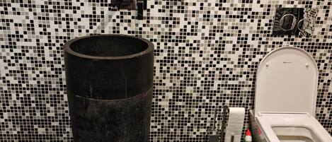 A bathroom with a mosaic tile wall, a round mirror, a toilet, and a large black trash bin.