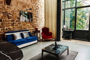 Una habitación con pared de piedra, un sofá azul, un sillón rojo y un ventanal con vistas a la vegetación.