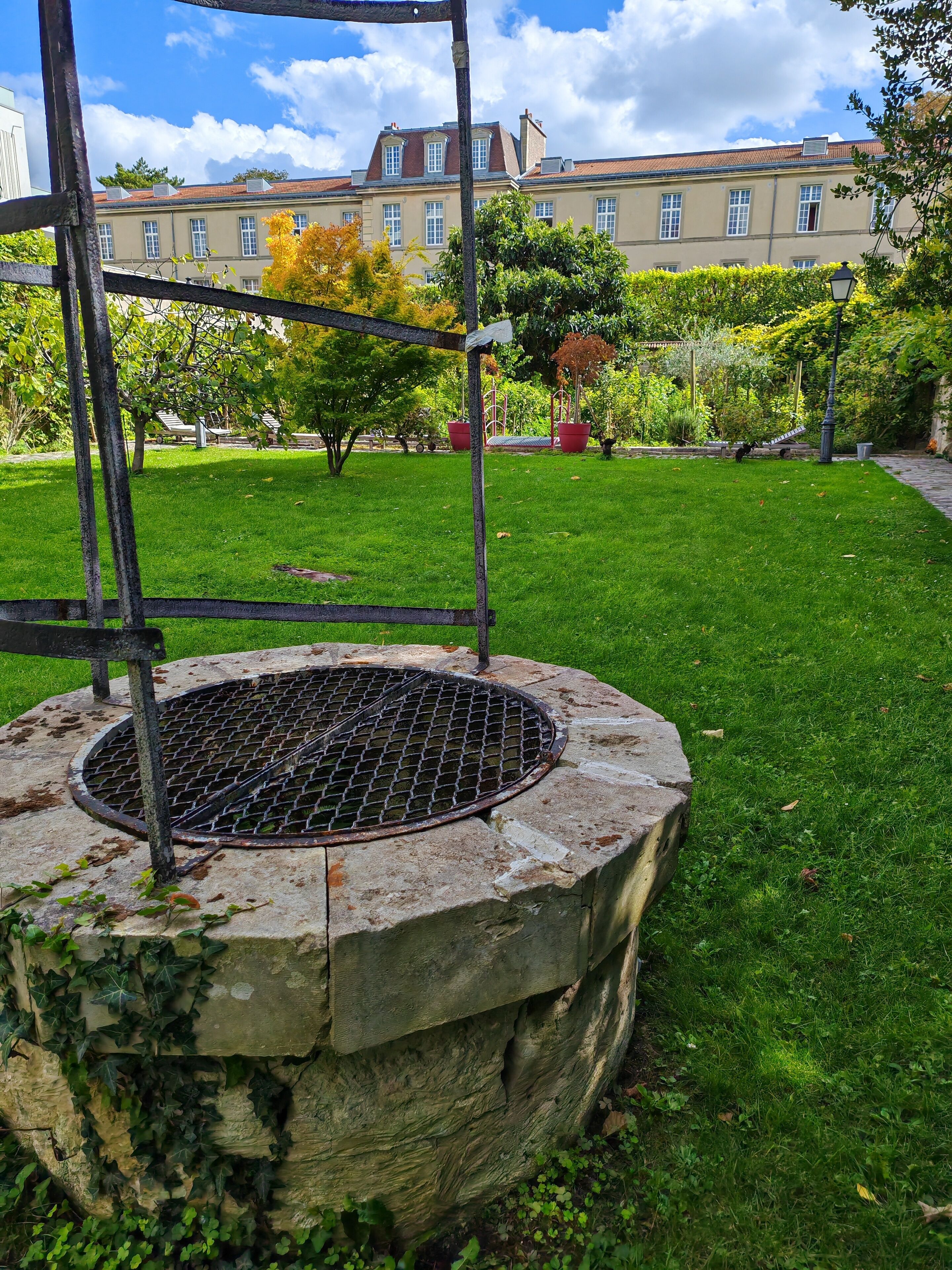Un puits circulaire en pierre, surmonté d’une grille métallique, entouré d’herbe et de lierre, situé dans un parc avec des arbres et un bâtiment en arrière-plan.