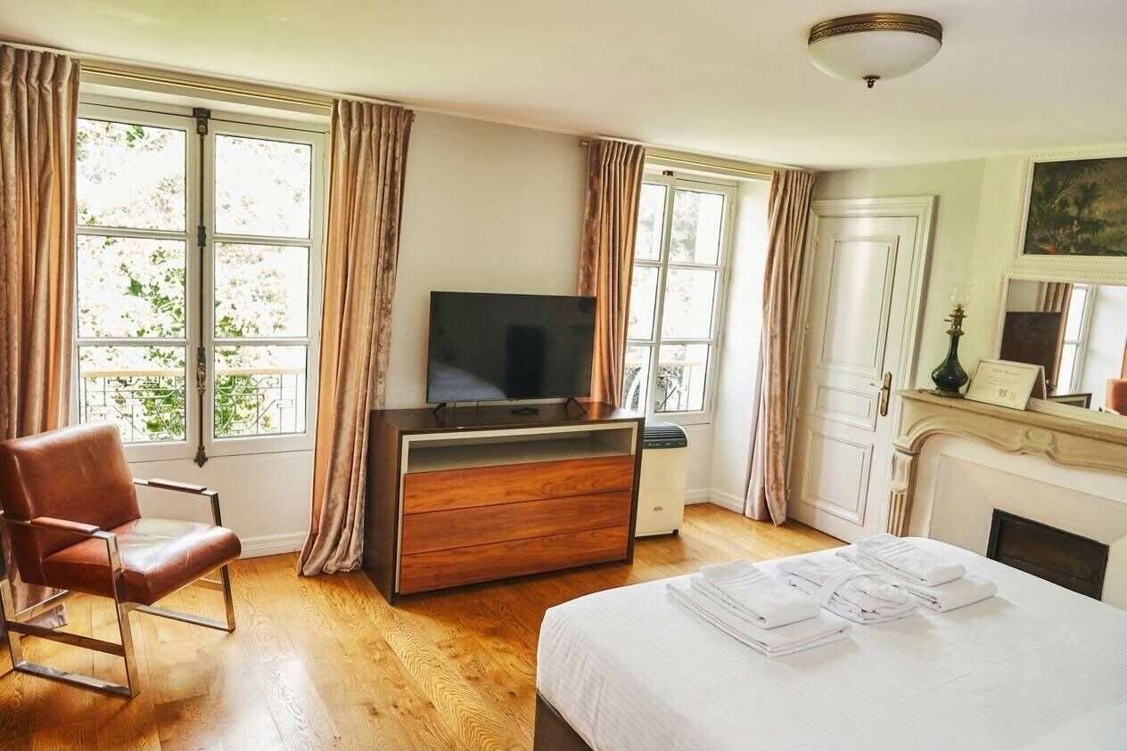 Une chambre d’hôtel avec un lit, un téléviseur posé sur une commode en bois, une chaise et de grandes fenêtres avec des rideaux.