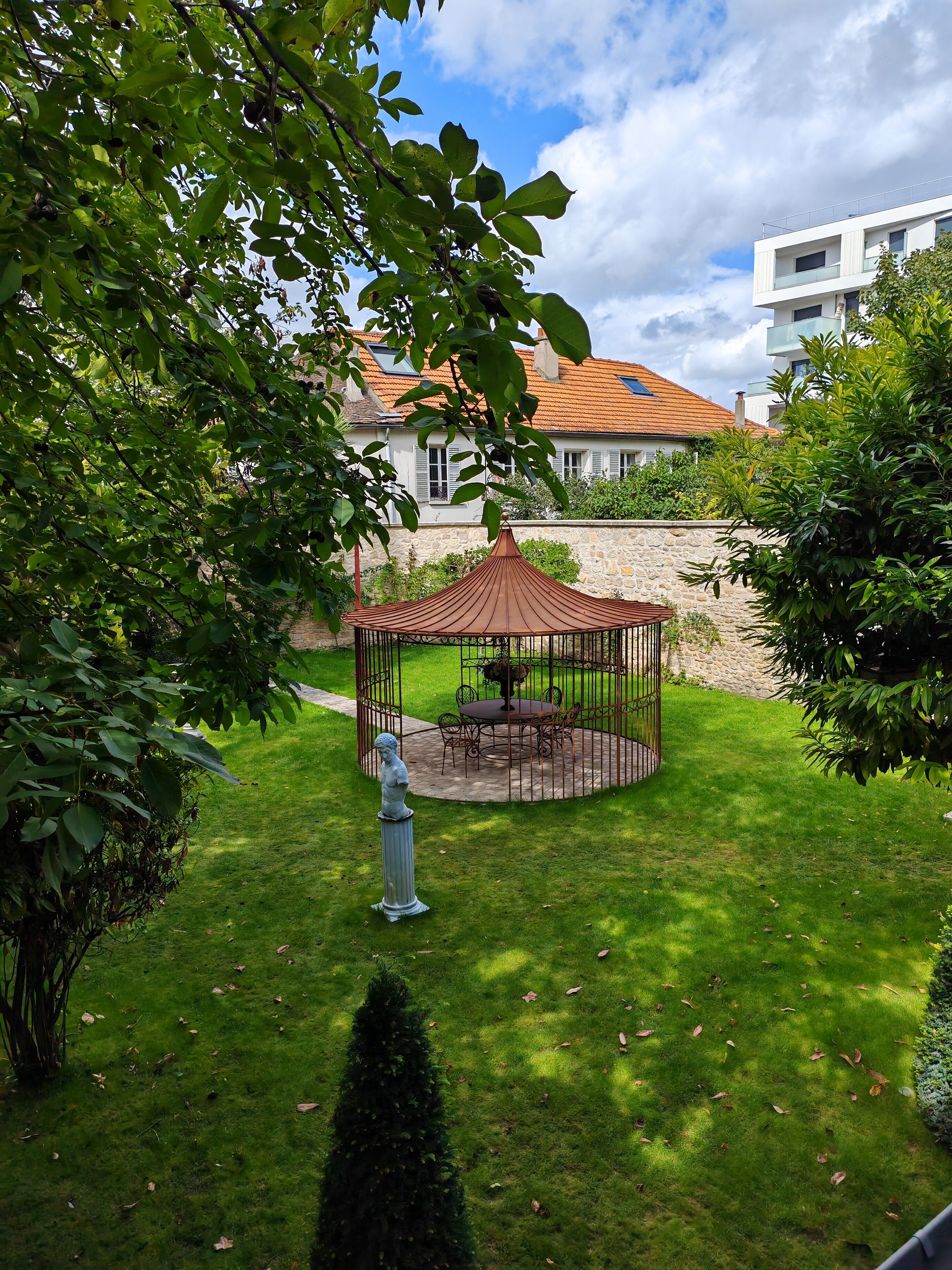 Un belvédère avec une table et des chaises, situé dans un jardin bien entretenu, entouré de verdure et délimité par un mur de pierres.
