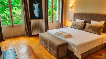 A bedroom with a large bed, a bench, a radiator, and a window with a view of greenery.