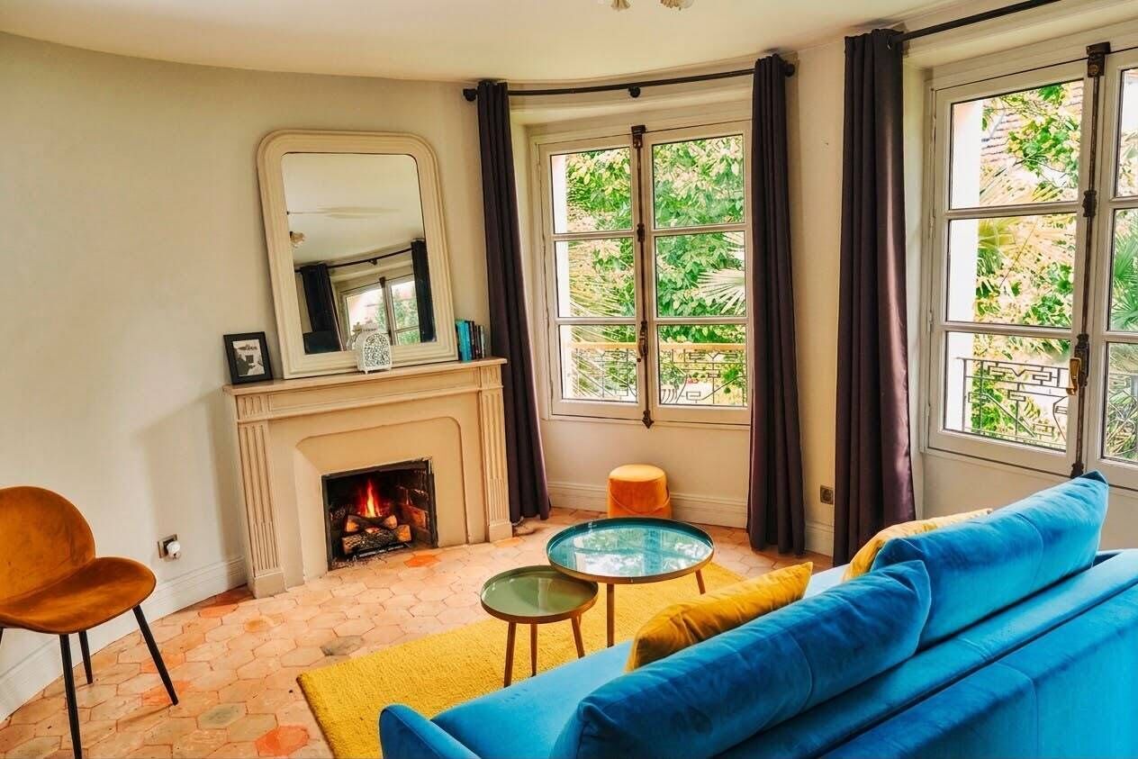 A living room with a fireplace, a blue sofa, a round glass table, and a view of greenery through large windows.