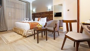 Habitación de hotel con cama, dos sillas, una mesita con fruta, televisión y un ventanal con cortinas.