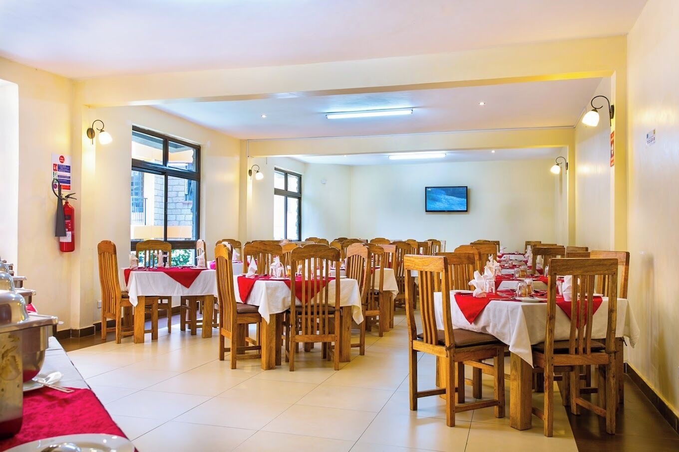 A dining area with multiple tables and chairs, a fire extinguisher on the wall, and a television mounted on the wall.