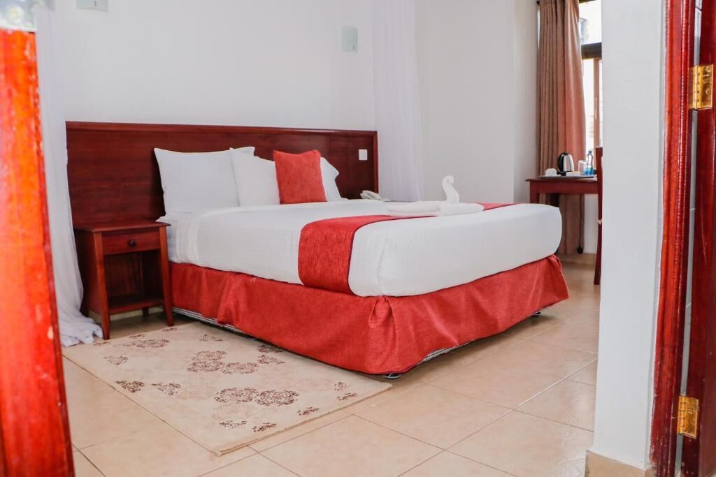 Habitación de hotel con una cama grande, cabecera de madera, mesita de noche, camino de cama rojo y una pequeña mesa con hervidor y tazas.