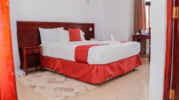 Habitación de hotel con una cama grande, cabecera de madera, mesita de noche, camino de cama rojo y una pequeña mesa con hervidor y tazas.