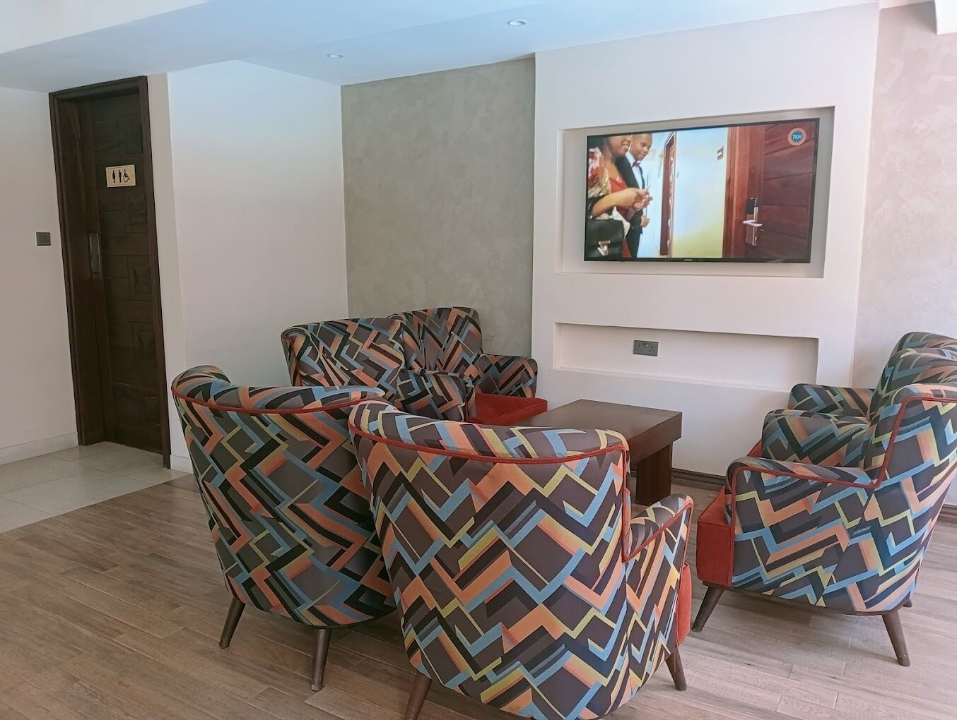 A modern living room with patterned armchairs, a small table, and a flat-screen TV mounted on the wall.