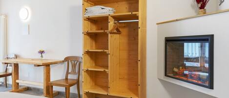 Una habitación con un armario de madera, una mesa con sillas, una chimenea y un televisor instalado en la pared.