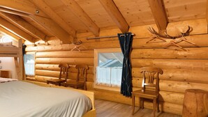 Una habitación tipo cabaña de madera con una cama, dos sillas, un ventanal con cortinas y cuernos de ciervo en la pared.