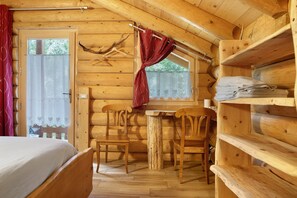 A wooden cabin interior with a bed, two chairs, a table, and a window with curtains.