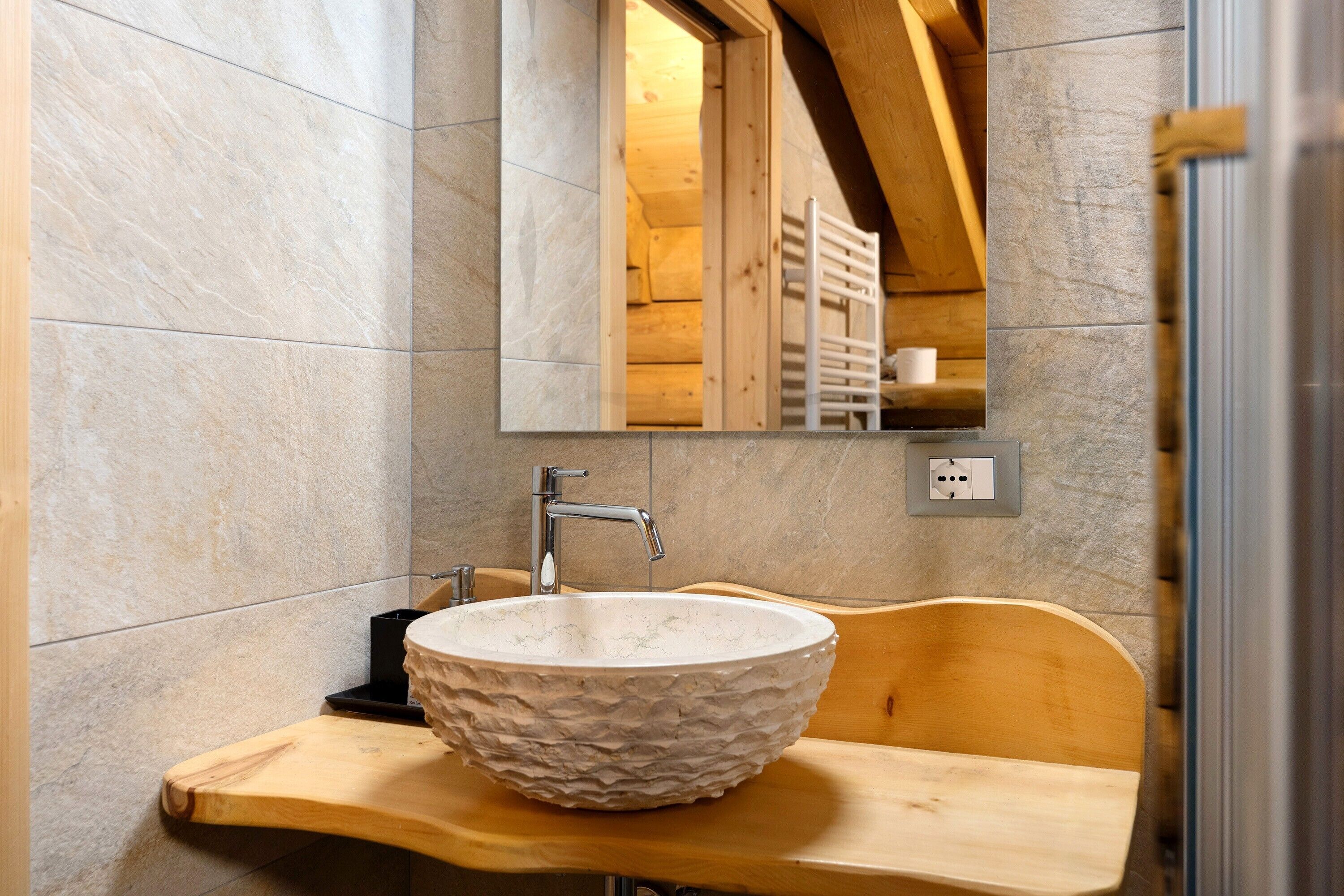 A bathroom with a stone wall, a wooden bench, a round sink, and a mirror reflecting a wooden ceiling and a towel rack.