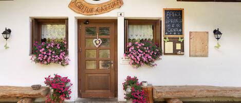 Un edificio con puerta de madera, dos cestas de flores, un banco y un cartel de pizarra.