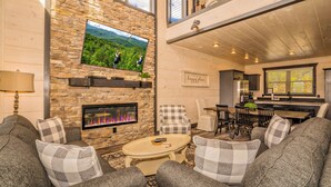 A cozy living room with a stone fireplace, a flat-screen TV mounted on the wall, and a dining area with a kitchen in the background. - Fantastic View|Theater|Indr Pool|Pool Table|HotTub (Sevierville)