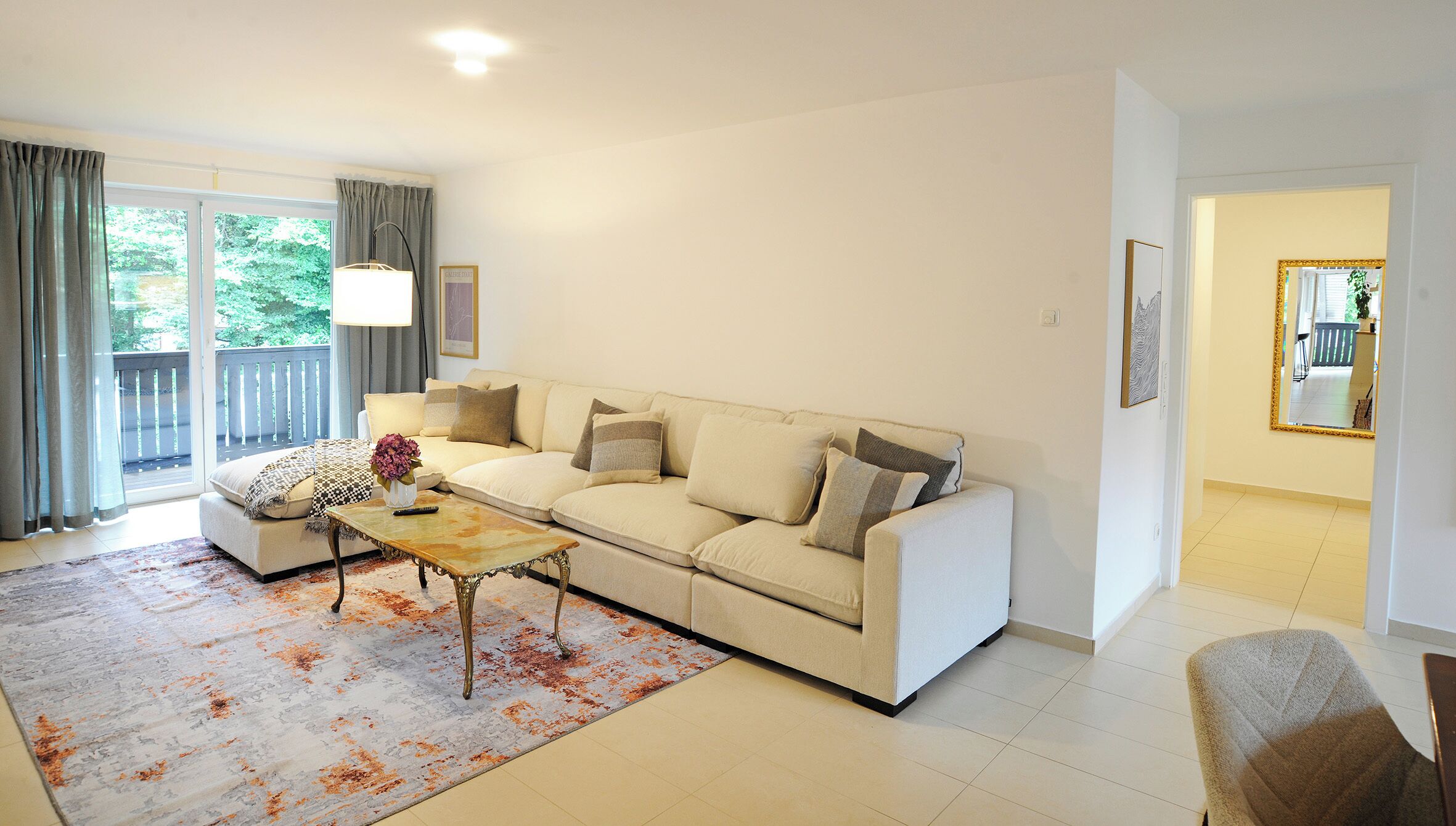 A living room with a beige sofa, a coffee table, and a patterned rug. There is a balcony with curtains and a view of greenery outside.