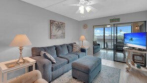 Un salon avec un canapé gris, un pouf et un téléviseur posé sur un meuble. On y trouve également un ventilateur de plafond, un tableau et une horloge au mur.