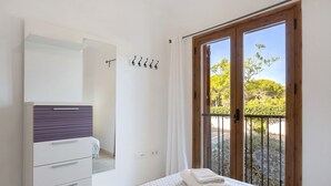 A bedroom with a bed, a wardrobe, a window with a view of greenery, and a balcony with a railing.