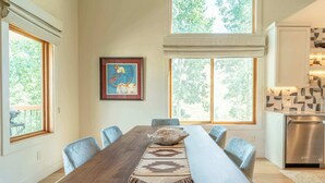 A dining area with a long wooden table, chairs, and a decorative rug. There are large windows with a view of trees. - Oak at Outrun, brand new remodel, fireplace! (Mt. Crested Butte)