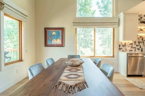 A dining area with a long wooden table, chairs, and a decorative rug. There are large windows with a view of trees. - Oak at Outrun, brand new remodel, fireplace! (Mt. Crested Butte)