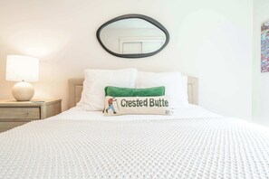 A neatly made bed with a white comforter, a green pillow with a character illustration, a round mirror, and a lamp on a bedside table. - Oak at Outrun, brand new remodel, fireplace! (Mt. Crested Butte)