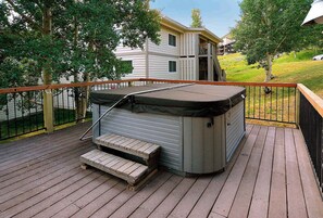 A hot tub on a wooden deck with a metal railing, surrounded by trees and a residential building in the background. - Oak at Outrun, brand new remodel, fireplace! (Mt. Crested Butte)