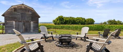A rustic outdoor seating area with Adirondack chairs and a fire pit, set against a backdrop of a wooden barn and a field.