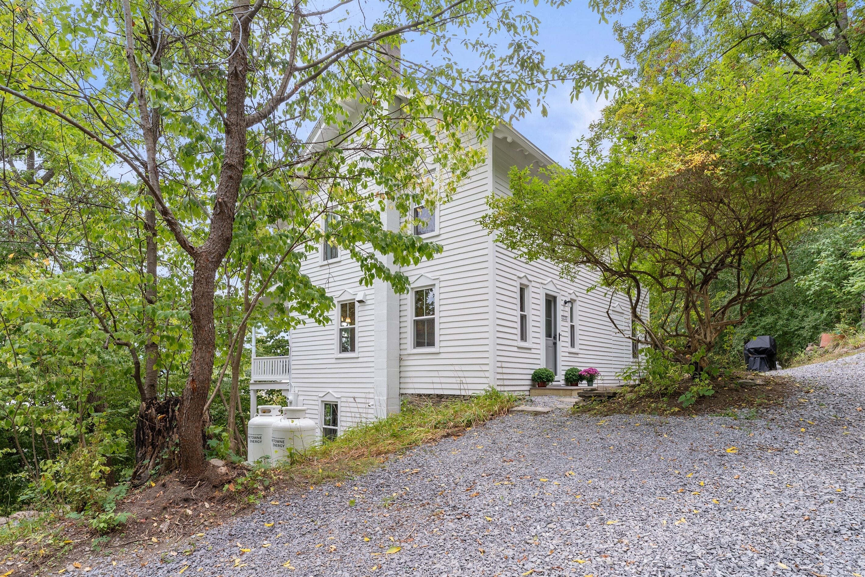 Une maison blanche avec une allée en gravier, entourée d’arbres et de verdure.