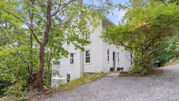 Une maison blanche avec une allée en gravier, entourée d’arbres et de verdure.