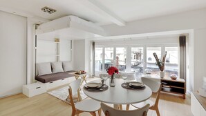 A modern, well-lit room with a dining area, a sofa with storage, and large windows offering a view of the city. - Cozy Studio - 2P - Place de la Bourse (Paris)